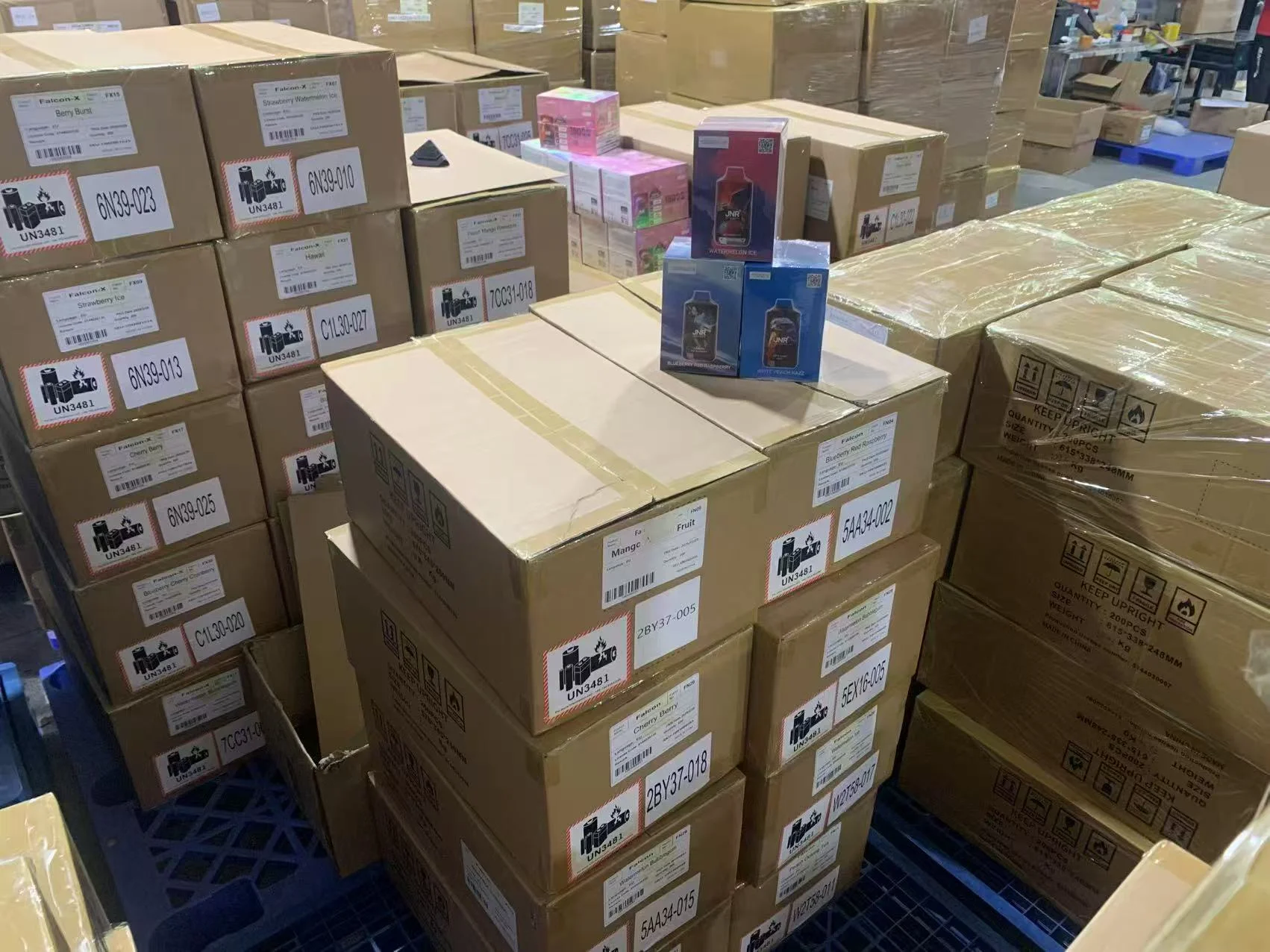 Stacks of cardboard boxes with electronic cigarette labels in a warehouse. Smaller product boxes visible on top.
