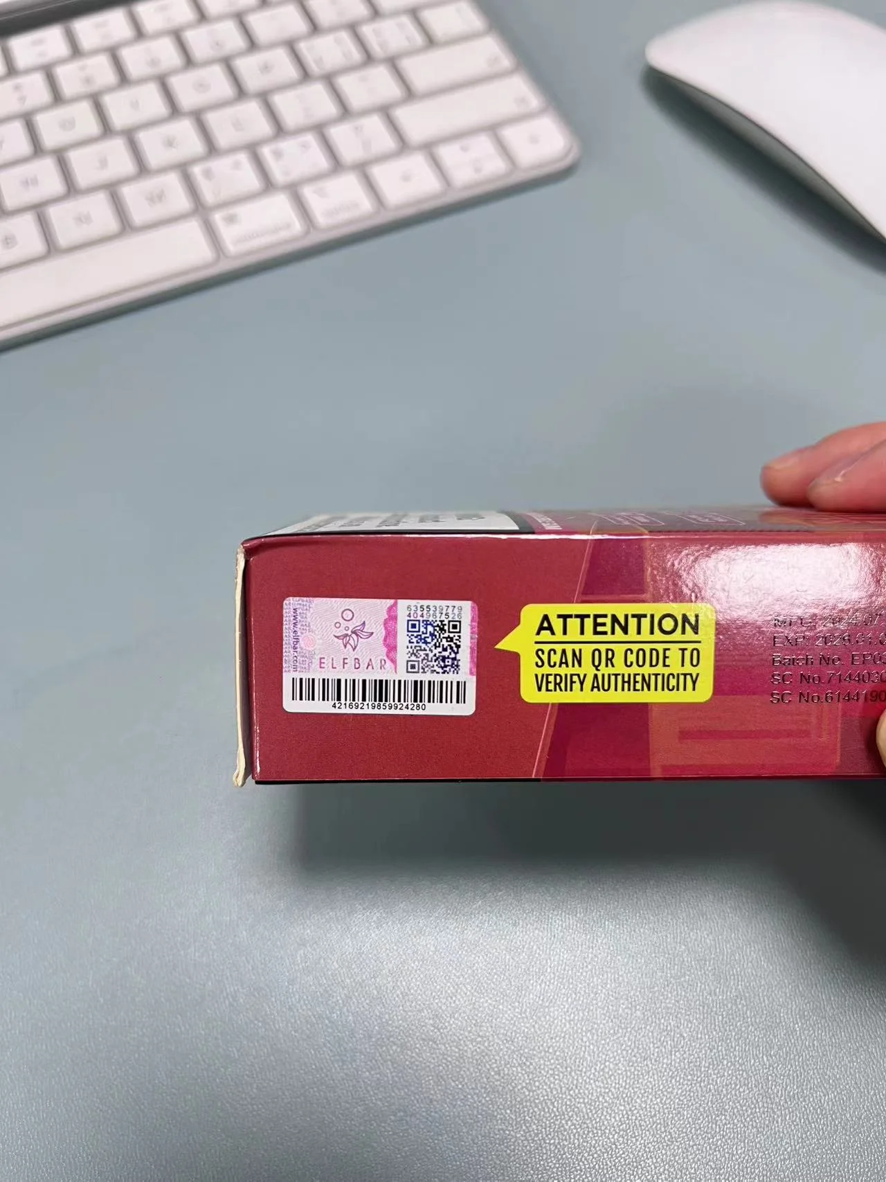 Red vape box with a QR code and yellow label instructing to scan for authenticity verification on a desk near a keyboard and mouse.