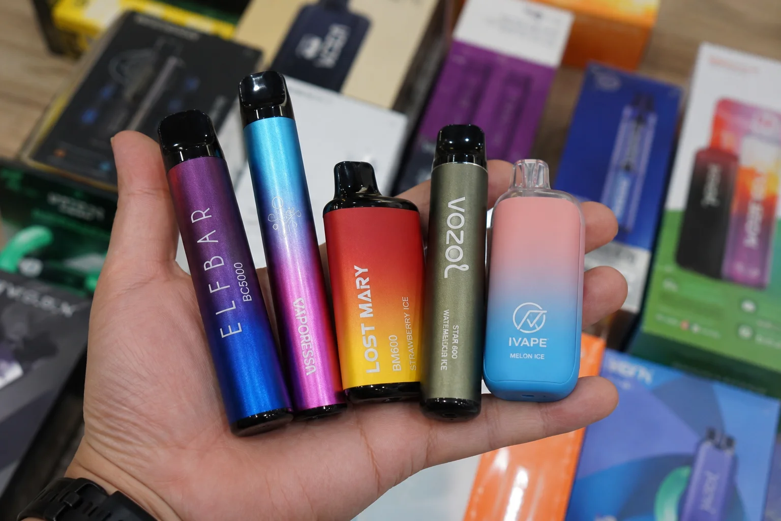 A hand holding five disposable vapes with vibrant designs from brands like Elf Bar, Lost Mary, Vozol, and Ivape, displayed among vape product packaging.