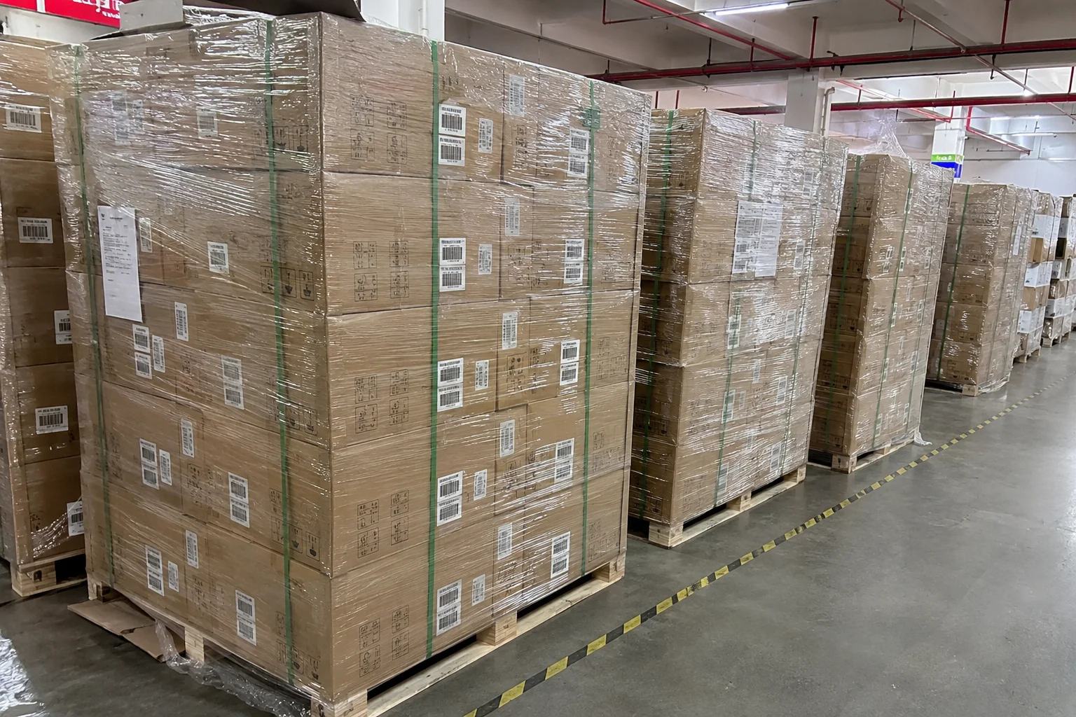 Stacks of cardboard boxes secured with plastic wrap and green straps on wooden pallets in a warehouse with bright lighting.